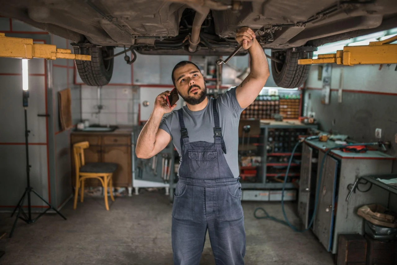 A mechanic working beneath a raised vehicle while holding a phone to his ear.